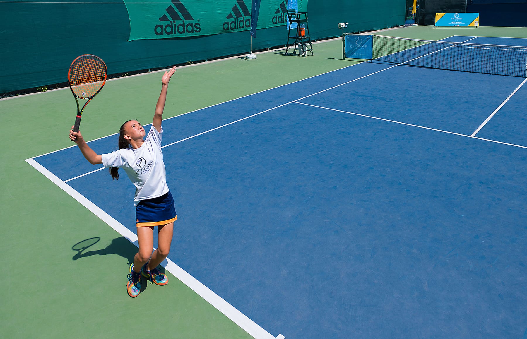 A CF Tennis Academy student serving on a blue hard court.